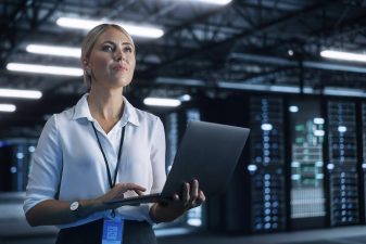 Nainen seisoo kannettava tietokone kädessään konesalissa.