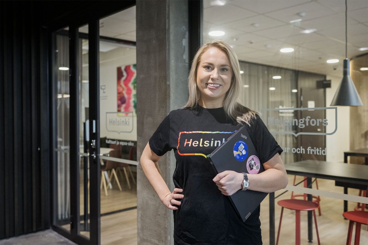 Nainen, joka on meidän asiakas ja Helsingin kaupungin työntekijä, hymyilee kannettava tietokone kainalossaan toimistolla.