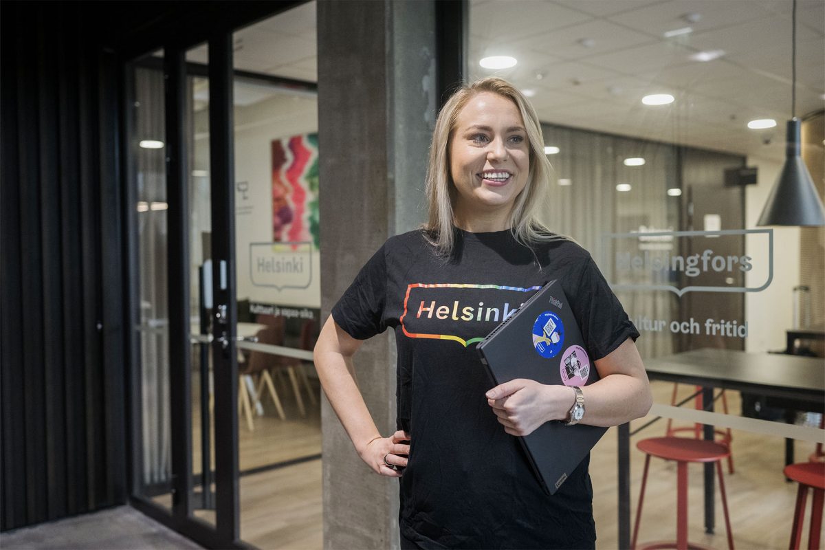 Nainen, joka on meidän asiakas ja Helsingin kaupungin työntekijä, hymyilee kannettava tietokone kainalossaan toimistolla.